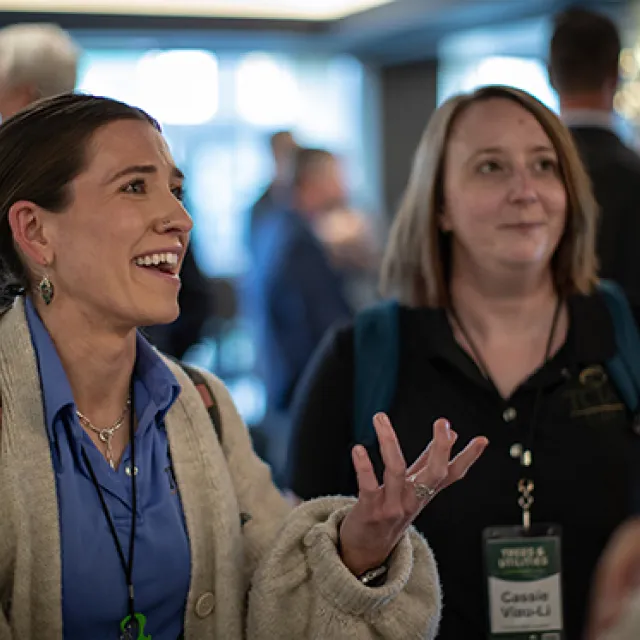 Two women engage in conversation at a professional networking event with other attendees blurred in the background.